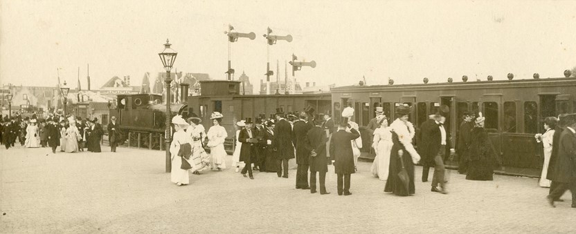 Een historisch afbeelding van een treinstation met wahtende passagiers.