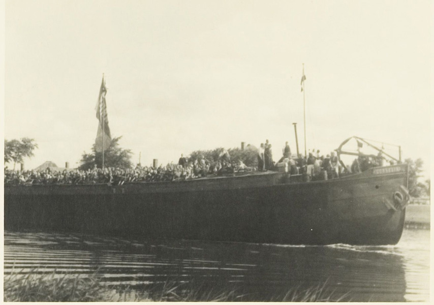 Het binnenvaartschip Cornelia met kinderen in het Noordhollands Kanaal ter hoogte van de vlotbrug bij Koedijk.  De kinderen keren terug uit Friesland, Groningen en Drenthe waar ze tijdens de barre laatste maanden van de bezetting als evacués onder waren gebracht. In totaal betrof het zo
